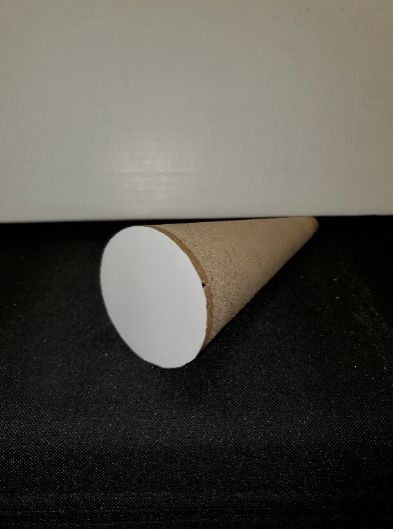 The cone lying with its long, curved surface along the table. The point of view is slightly above the plane of the table. The circular base of the cone is closest to the viewer and is fully visible; a circular piece of chipboard has been cut to match the base and glued to it. The cone tapers away from the viewer at about a 45 degree angle towards the right. Because the cone tapers away from the viewer at an angle, the two-dimensional aspect of the circular base is somewhat distorted into an oval shape.