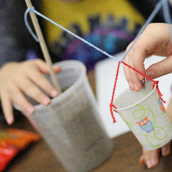 A cup of washers suspended on a string attached to two wooden dowels standing in cups of soil.