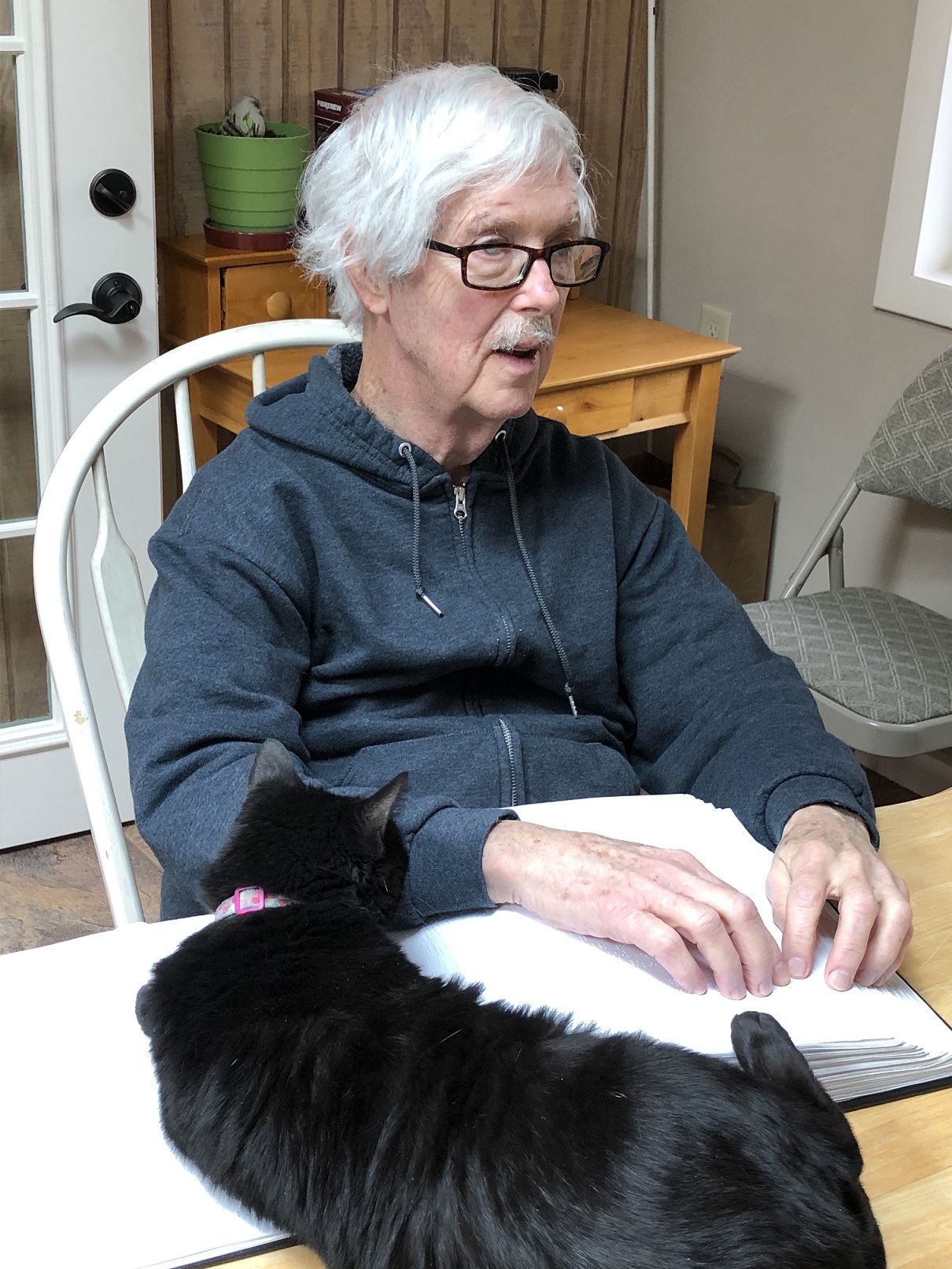 Bob Gardner reads from a Braille book with his cat Gracie on the table next to him.