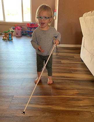 Kadyn standing in our family room holding his cane and smiling at the camera.