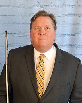 Everette Bacon wearing a black blazer with a cream colored shirt and yellow tie.
