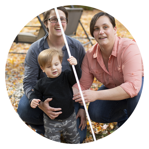 Moms play in the leaves with their young son. He holds his mom's long white cane.