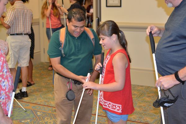 A blind man helps a blind girl use her long white cane.