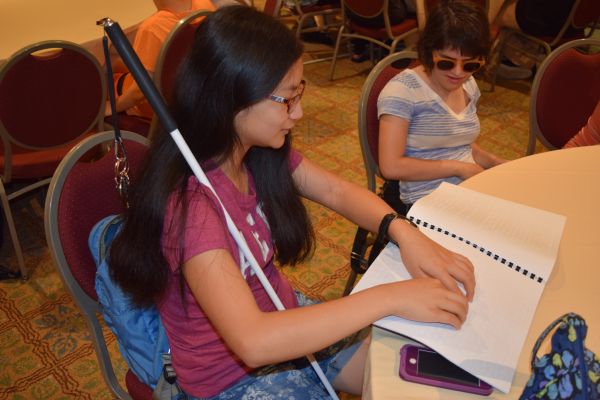 A girl with a white cane sits at a table and reads from a Braille book.