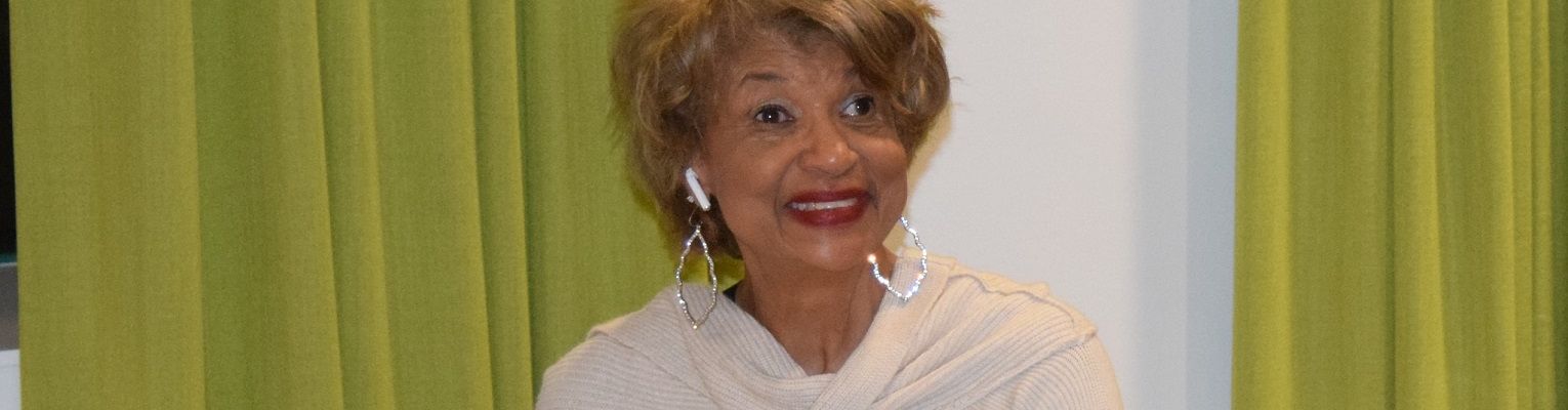 Ever Lee Hairston smiles as she sits by a piano during an NFB National Convention.