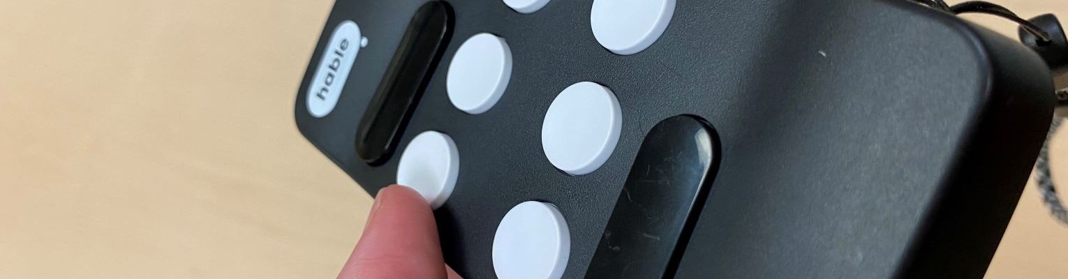 Hable One Braille Input in palm of hand with thumb near raise button. The Hable One is a black rectangle with 6 round white buttons, two (one above and one below the white round buttons) black rectangle buttons rounded on the ends, and a black and white cord wrist strap.