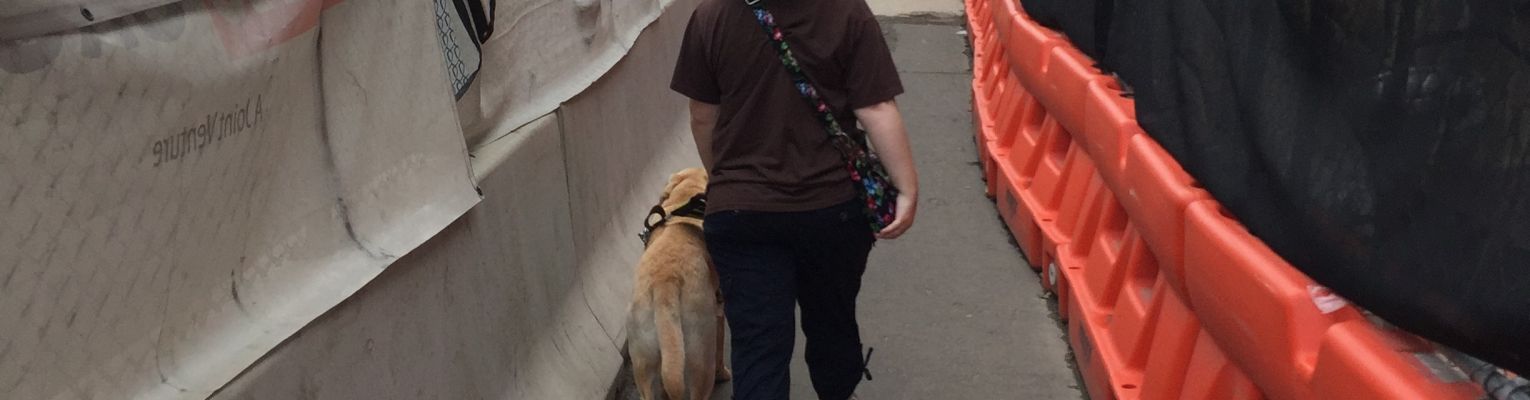 Ashley and Fuller navigating a pedestrian path made in the midst of heavy construction in Washington DC; an orange barricade is on their right with a concrete barricade on the left; buildings are in view beyond the barriers on the right and in the distance.