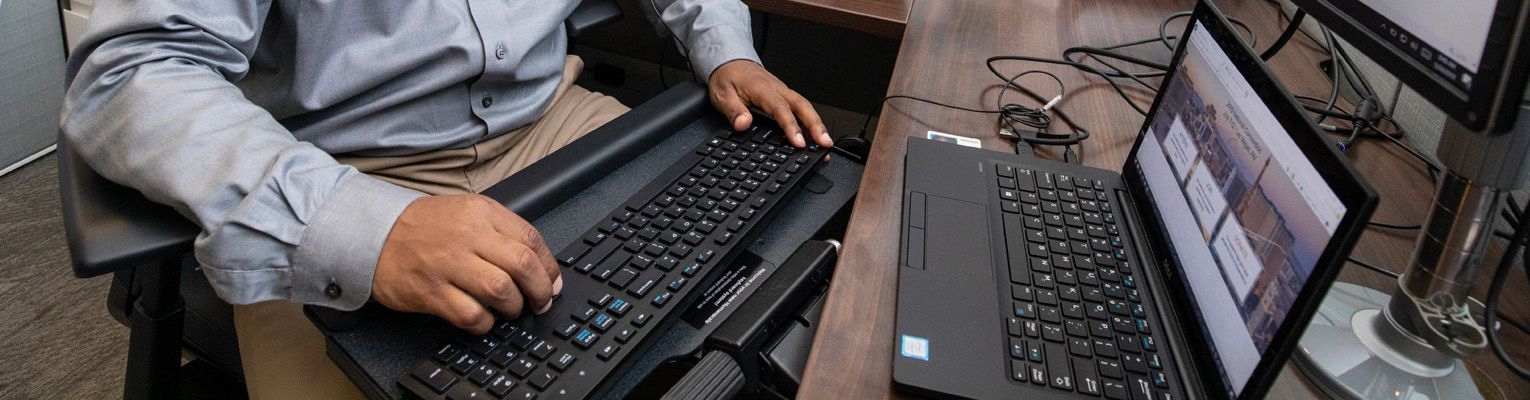 Person typing on a keyboard on their lap with a laptop sitting on the table in front of them. The laptop display shows an open website.