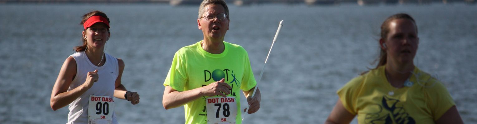 A blind man runs a road race; he is using a long white cane.