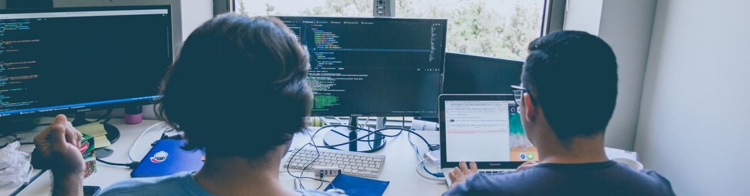 Two men sit at a desk facing computer screens with code on them.