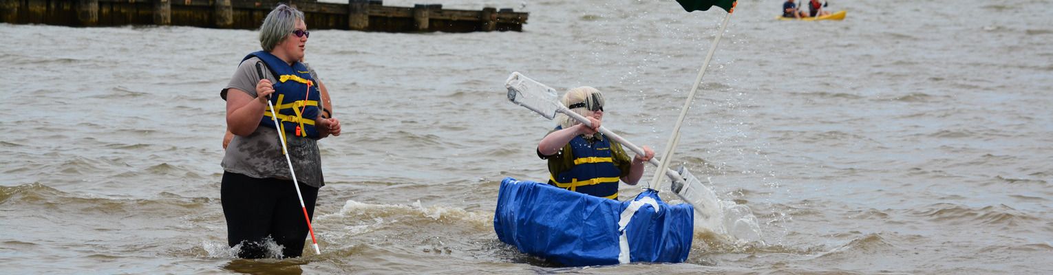 A STEM EQ participant paddles the boat their team built in the lake while another student stands nearby.
