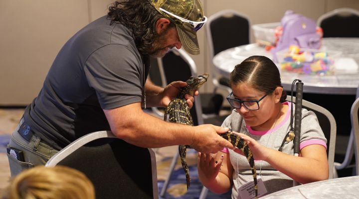An alligator trainer holds a small alligator as a blind child gently pats it